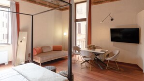 Moderni hotellihuone, jossa on sänky, sohva, työpöytä tuoleineen, taulutelevisio ja suuri ikkuna, jossa on verhot.