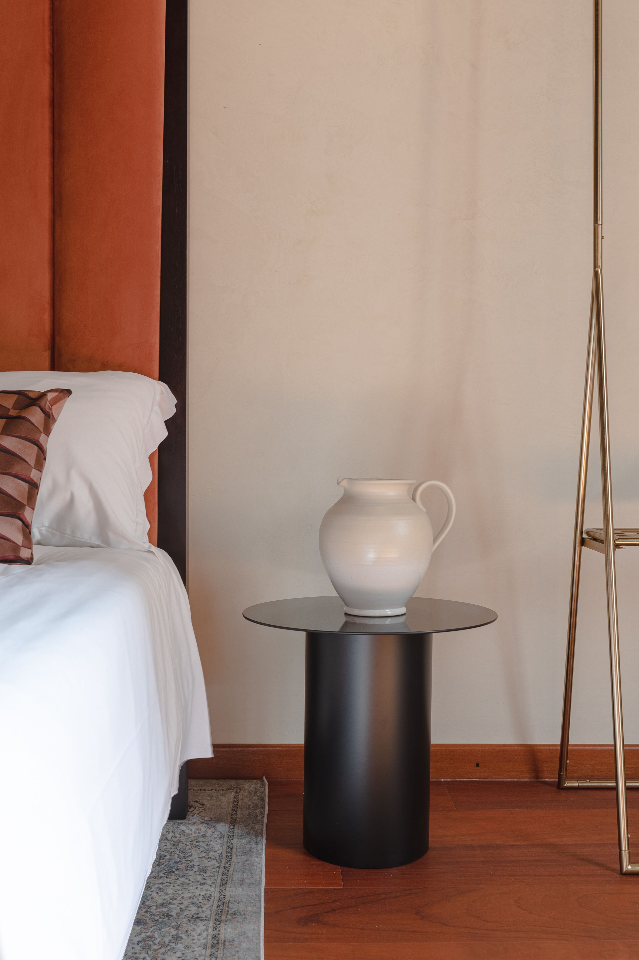 Une chambre d’hôtel avec un lit, une petite table sur laquelle se trouve un pichet blanc, et un lampadaire.