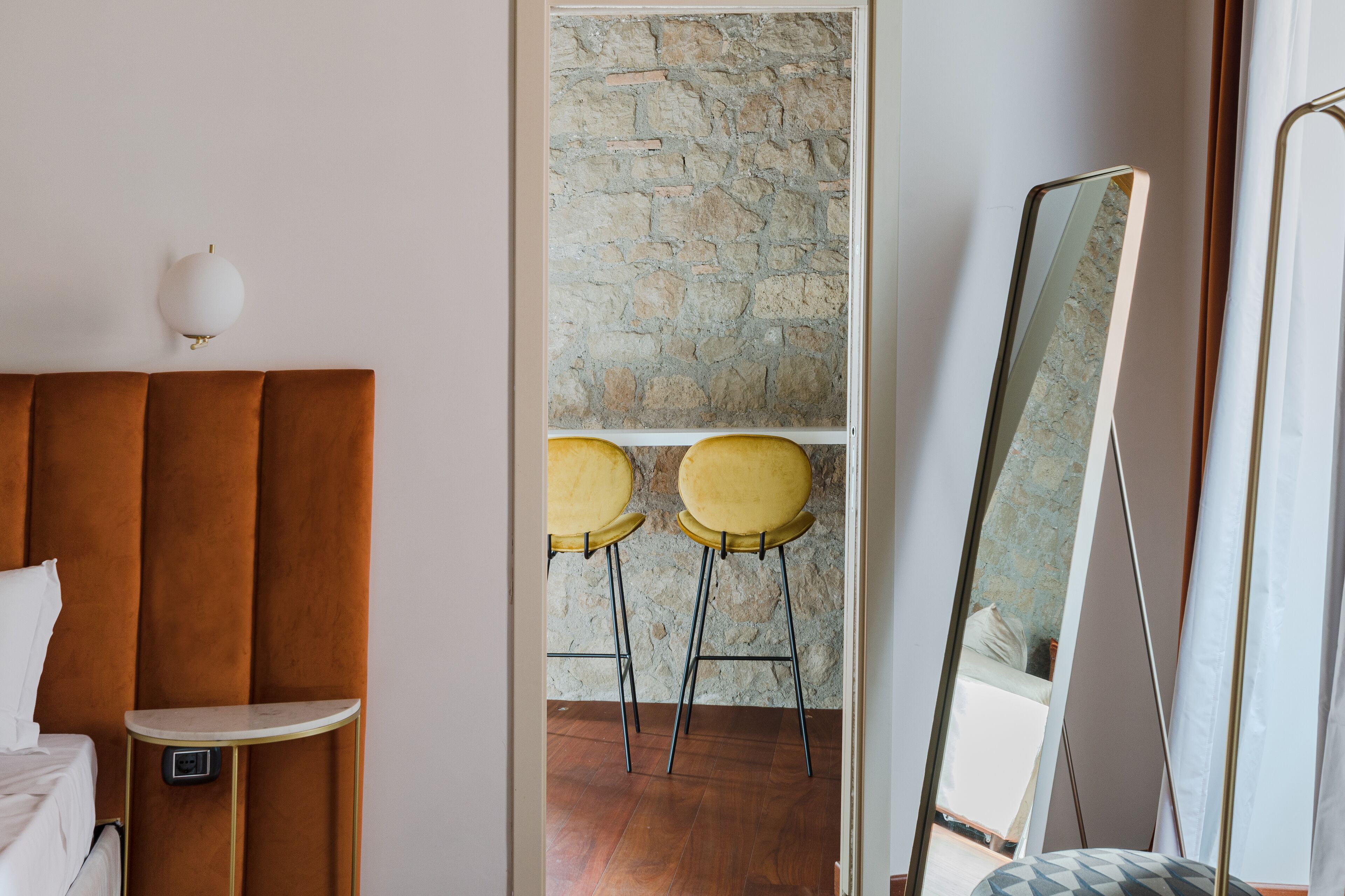 A bedroom with a brown headboard, a bedside table with a lamp, two yellow chairs, a stone wall, and a large mirror.