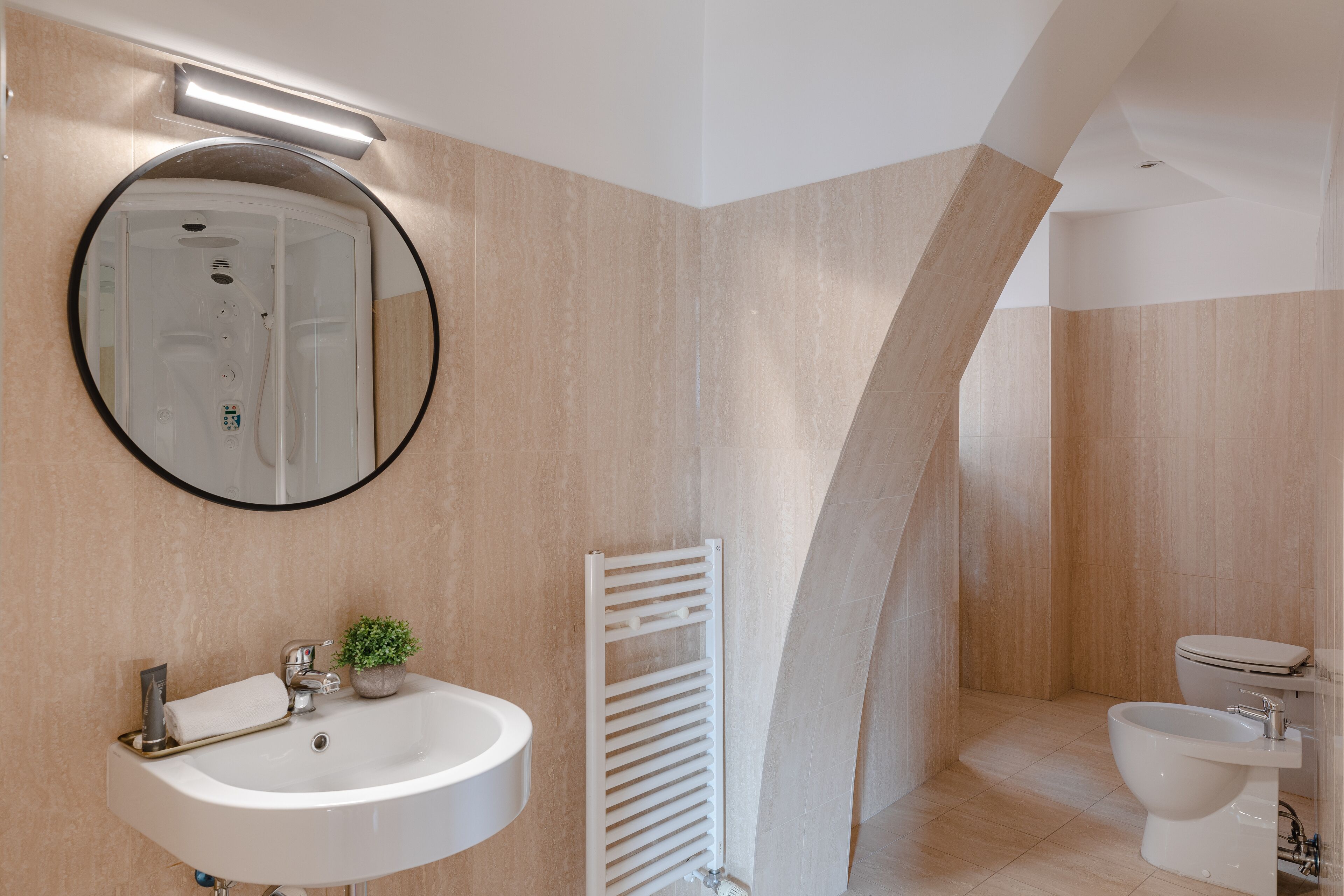 Un baño moderno con un espejo redondo, un lavabo, un perchero para toallas, un radiador y un bidé.