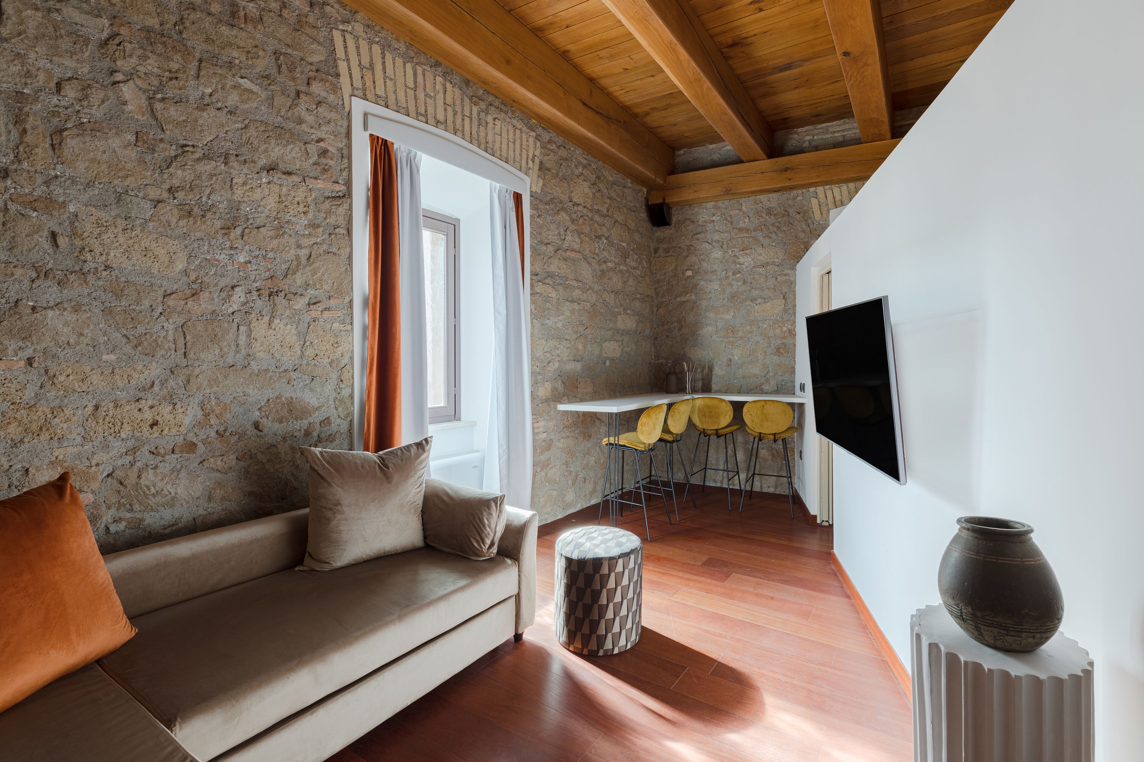 Un salon chaleureux avec un mur en pierre, des poutres en bois, un canapé, un pouf rond, un coin repas avec des chaises jaunes et un téléviseur fixé au mur.