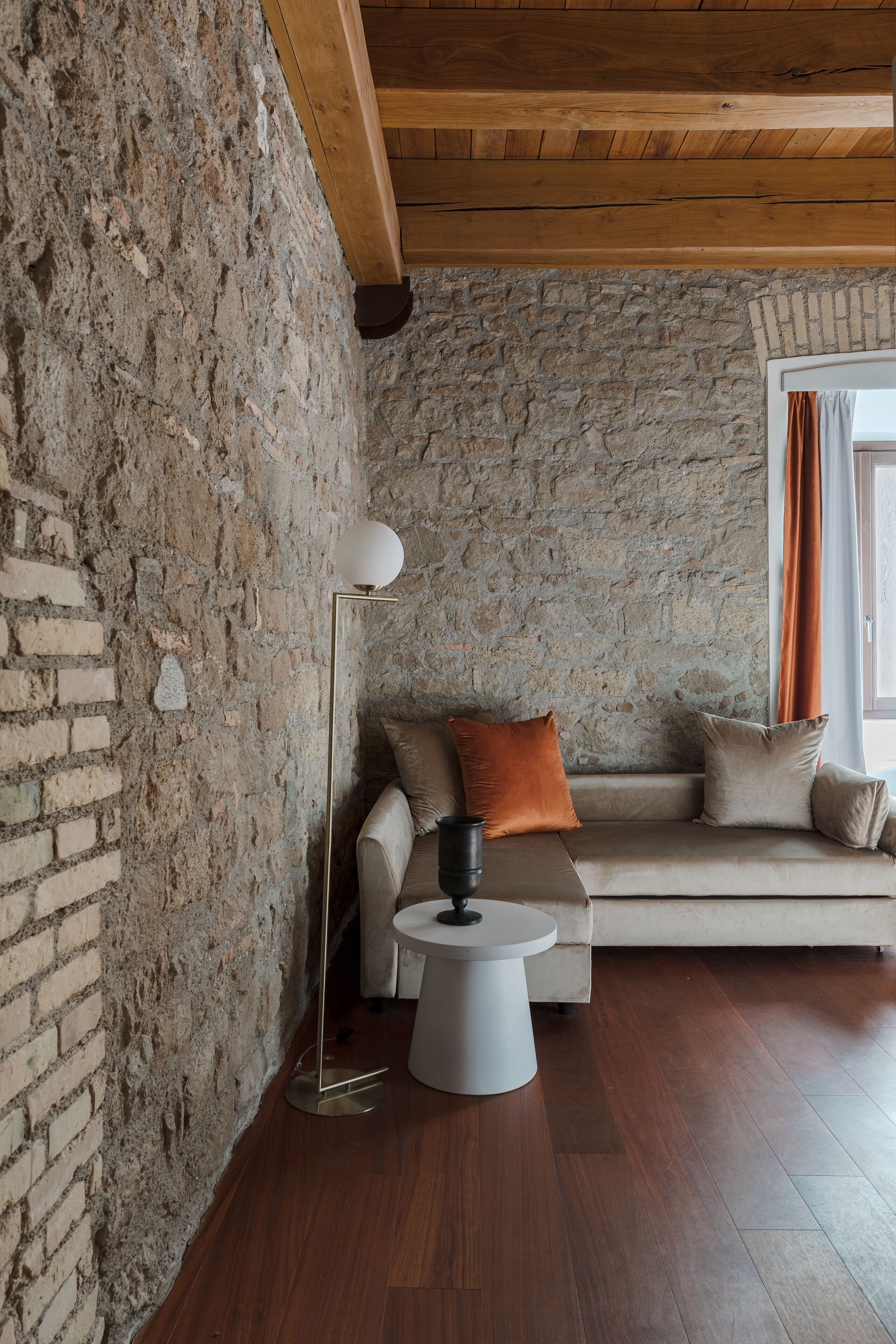 Une pièce chaleureuse avec un mur en pierre, un plafond en bois, un canapé avec des coussins, une lampe sur pied et une petite table ronde.