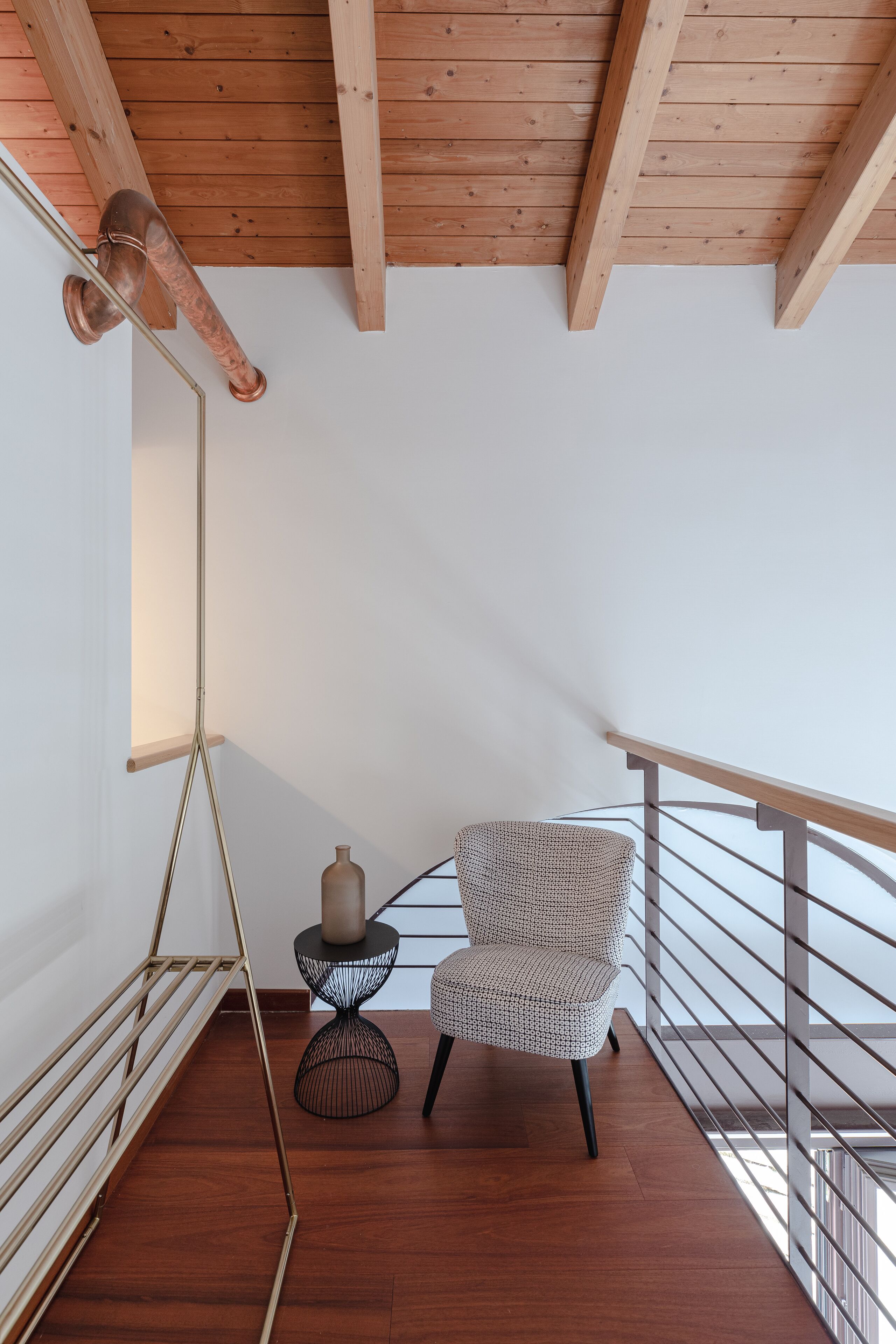 A modern room with a wooden ceiling, a metal floor lamp, a patterned armchair, a small round table with a vase, and a metal railing.