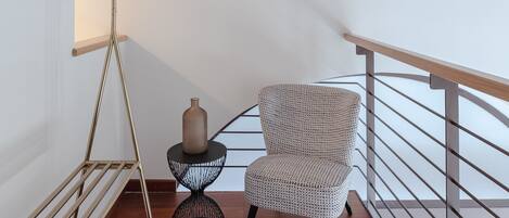 Una habitación moderna con techo de madera, lámpara de pie de metal, sillón estampado, una mesita redonda con un florero y barandilla de metal.