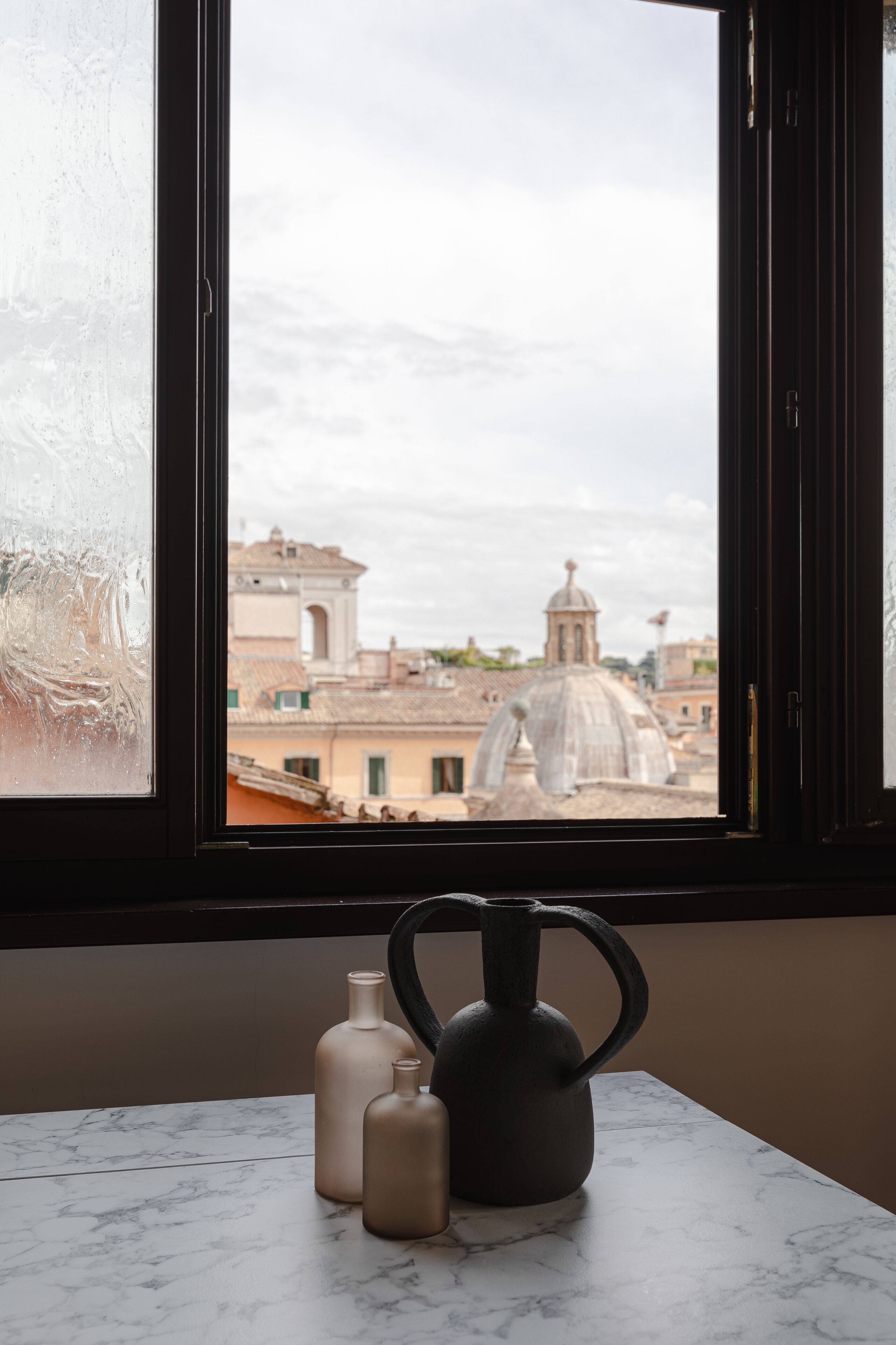 A window with a view of a cityscape, a marble table with three bottles, and a black kettle.