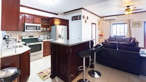 A kitchen with wooden cabinets, a refrigerator, a microwave, and a stove. There is a bar stool and a sofa in the living area.