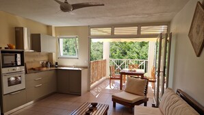 A compact kitchen with a microwave, oven, and sink, a wooden table with bowls, a sofa, and a balcony with a view of greenery. - Nice Duplex 6 persons in secured residence with swimming pool (Le Diamant)