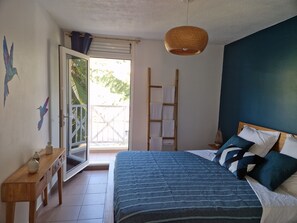 Een slaapkamer met een bed, een houten nachtkastje, een hanglamp, een raam met uitzicht op groen, en een deur naar een buitenruimte.