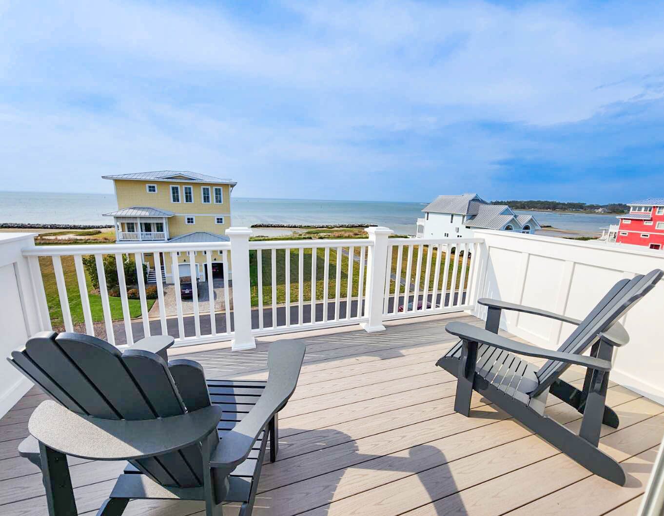 A deck with two Adirondack chairs and a table, overlooking a beach with houses and a clear sky.