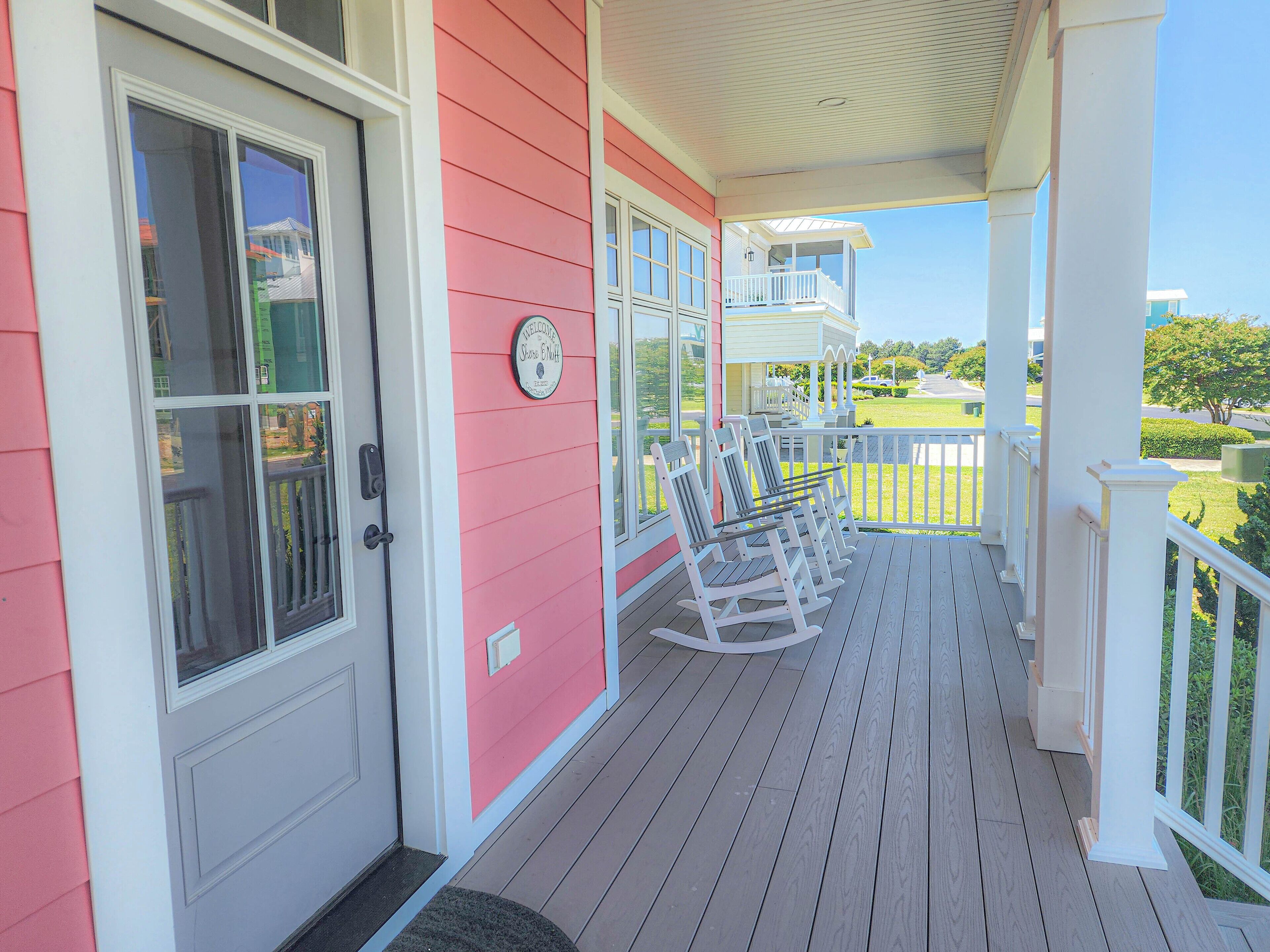 A porch with rocking chairs, a welcome sign, and a view of a green lawn and a building in the background.