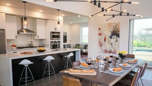 A modern kitchen with a central island, bar stools, and a dining area with a long table set for six. - Year-Round Heated Pool | Hot Tub | Fire Pit | Huge Lawn | Vineyard Views (Kenwood)