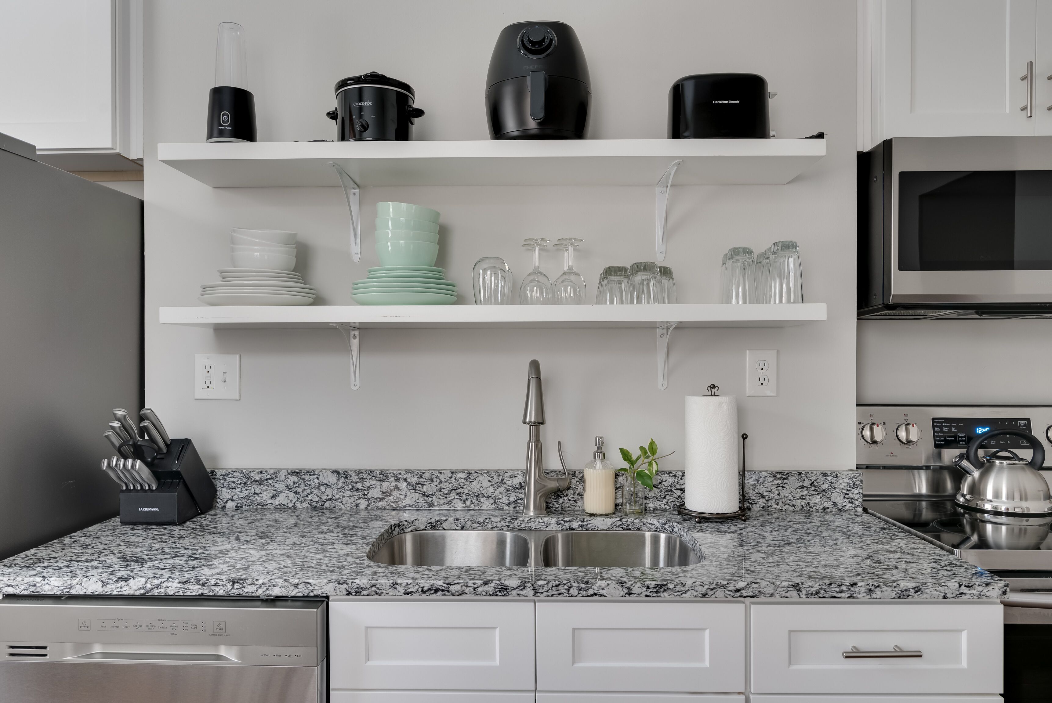 Cocina moderna con encimera de granito, pileta de acero inoxidable y muebles de cocina blancos. Se observan estantes sobre la encimera con utensilios y electrodomésticos.