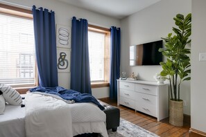 Un dormitorio con cama, cómoda, televisor y ventana con cortinas.