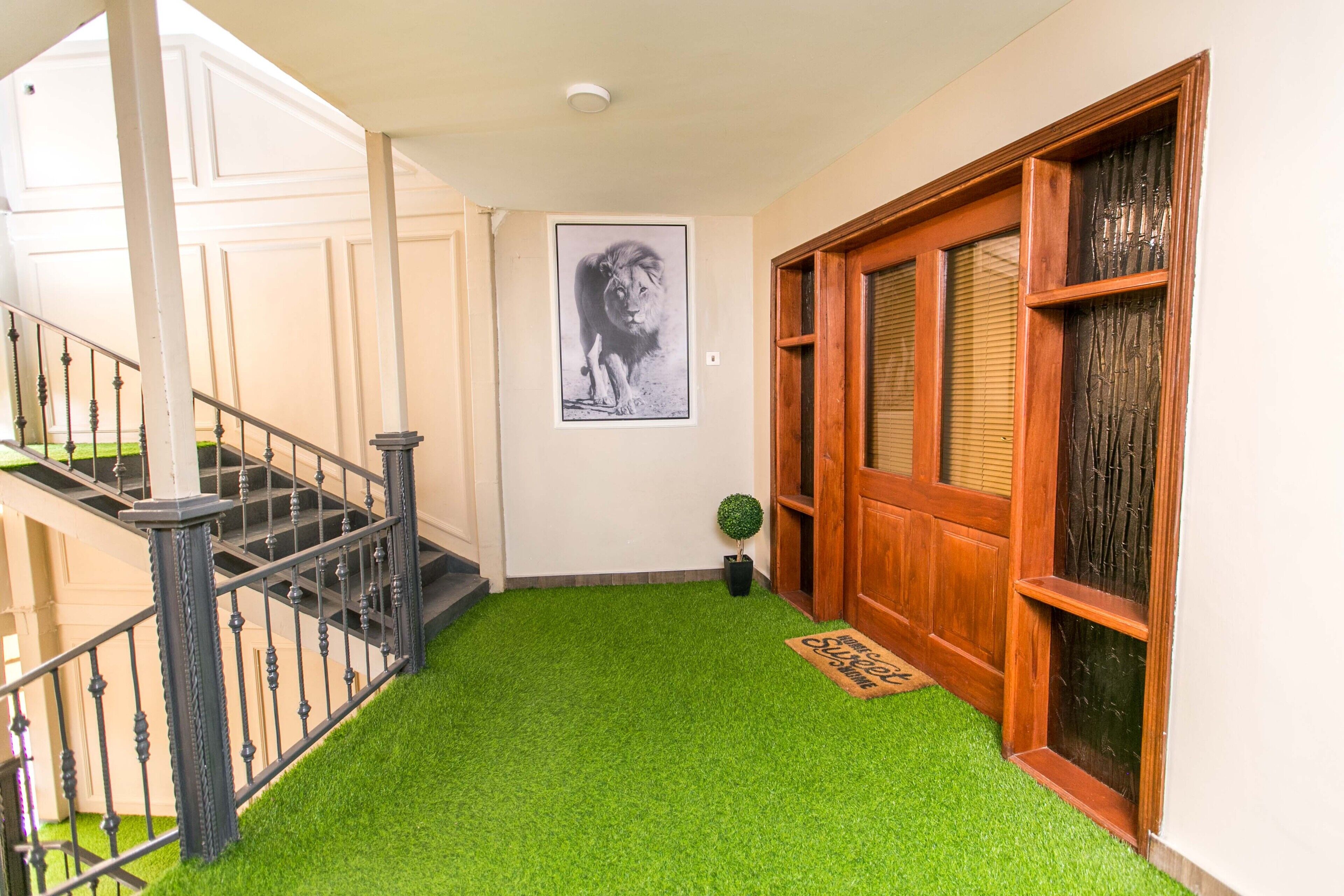 A hallway with artificial green carpet, a wooden door with glass panels, a black metal staircase with balusters, and a framed picture of a lion on the wall.