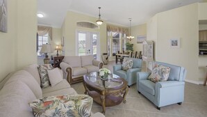 A spacious living room with a beige sofa, a blue armchair, and a glass coffee table. There is a dining area in the background with a table and chairs. - Traditional Inland Home | Pool/Spa/Steps to Shops (Marco Island)