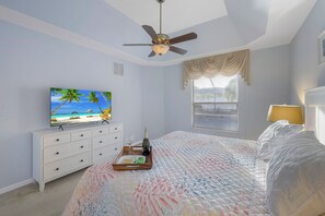 A bedroom with a bed, a ceiling fan, a TV on a dresser, a window with curtains, and a nightstand with a lamp.