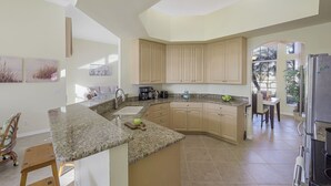 A modern kitchen with granite countertops, beige cabinetry, and a dining area with a table and chairs. - Traditional Inland Home | Pool/Spa/Steps to Shops (Marco Island)