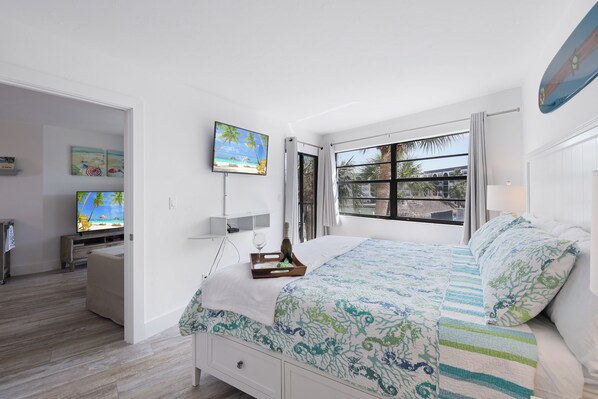 A bedroom with a bed, a TV, a window with curtains, and artwork on the wall.
