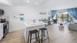 A modern kitchen with a central island, bar stools, and a dining area with a sofa and table. - Island-Style Riverfront Condo | Pools & Spas (Marco Island)