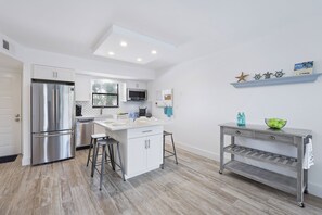 A modern kitchen with a stainless steel refrigerator, a microwave, and a sink. There is a bar stool and a small table with a bowl on it.