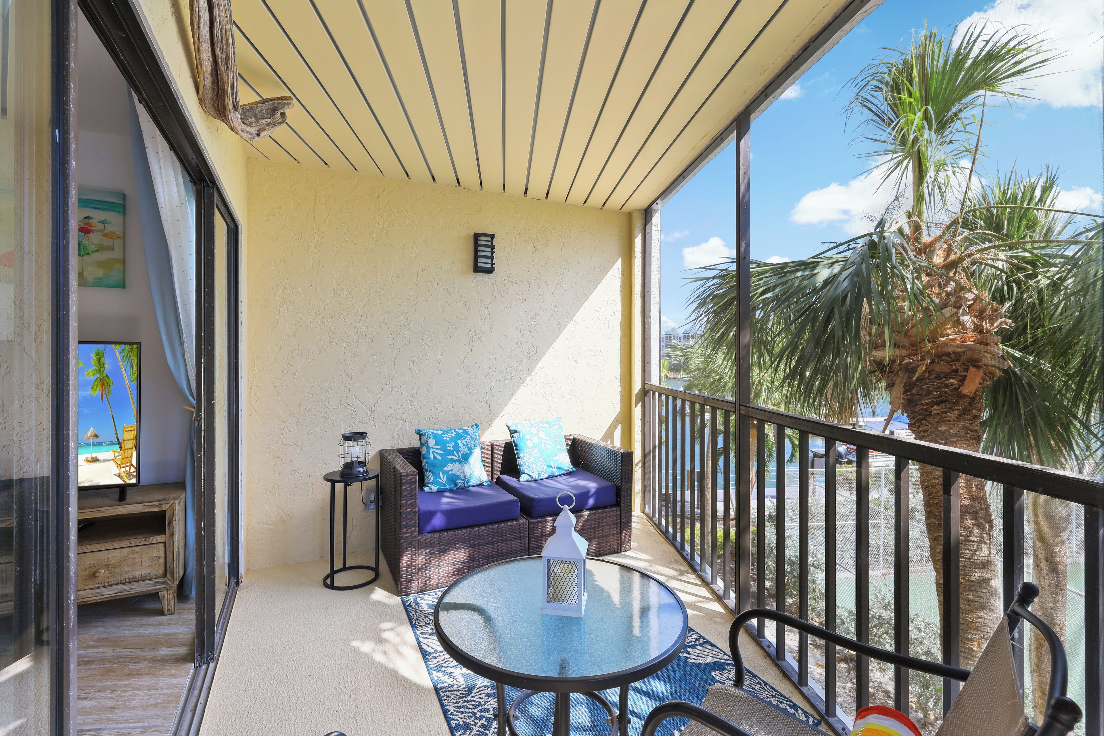 A balcony with wicker furniture, a glass table, and a view of palm trees.