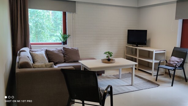 A living room with a sofa, a coffee table, a TV on a stand, a chair, and a window with a view of trees. - THE Roomz  (Kotka)