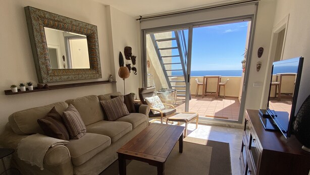 Ein Wohnzimmer mit Sofa, Couchtisch und Fernseher. Ein Balkon mit Tisch und Stühlen ist ebenfalls vorhanden.