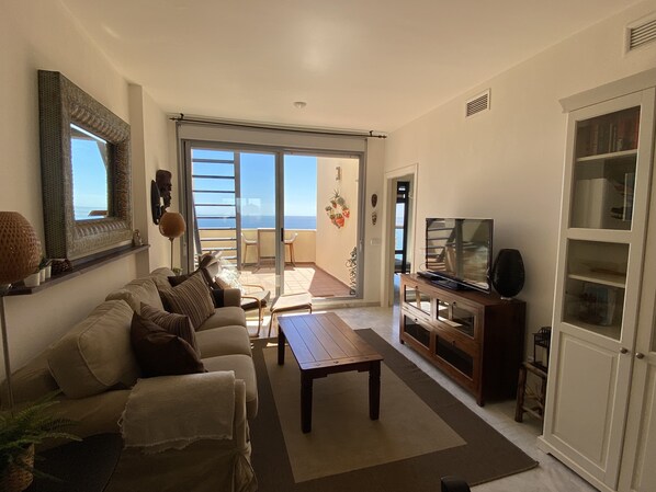 A living room with a sofa, coffee table, TV, and a sliding glass door leading to a balcony with a view of the sea.