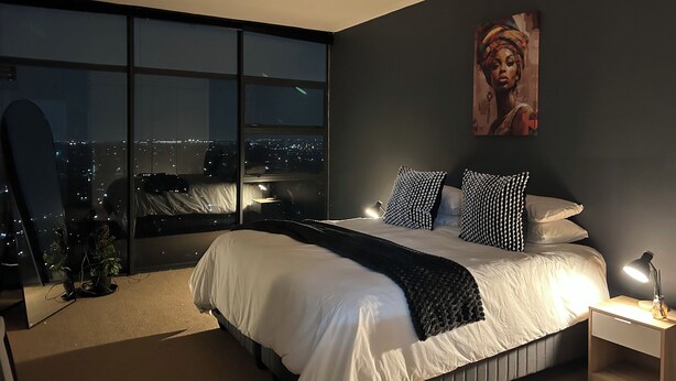 Een moderne slaapkamer met een groot bed, een nachtkastje, een bureau en een stadszicht door grote ramen.