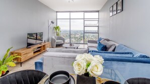 Een moderne woonkamer met een groot raam, een grijze bank, een blauwe bank, een televisie op een houten dressoir en een plant.