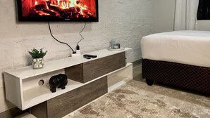 A modern bedroom with a TV mounted on a white brick wall, a wooden nightstand, a bed with a dark headboard, and a patterned rug.
