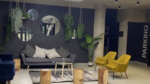 A modern lobby with a grey sofa, yellow chairs, and wooden stools. There are hanging plants and mirrors on the wall.