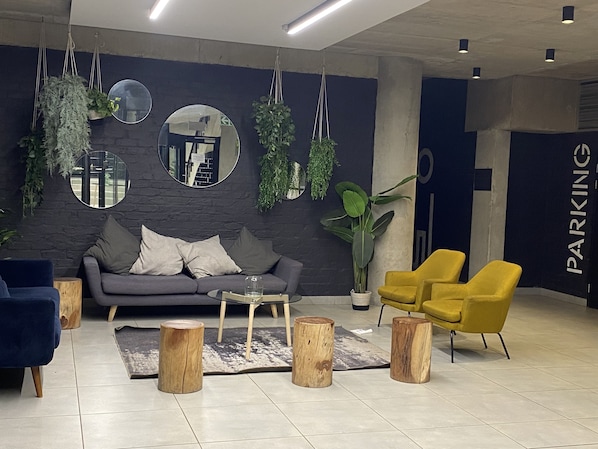 Un vestíbulo moderno con un sofá gris, sillas amarillas y taburetes de madera. Hay plantas colgantes y espejos en la pared.