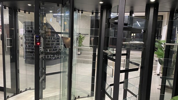 A revolving door with glass panels and a metal frame, leading to a lobby area with a staircase and a plant.