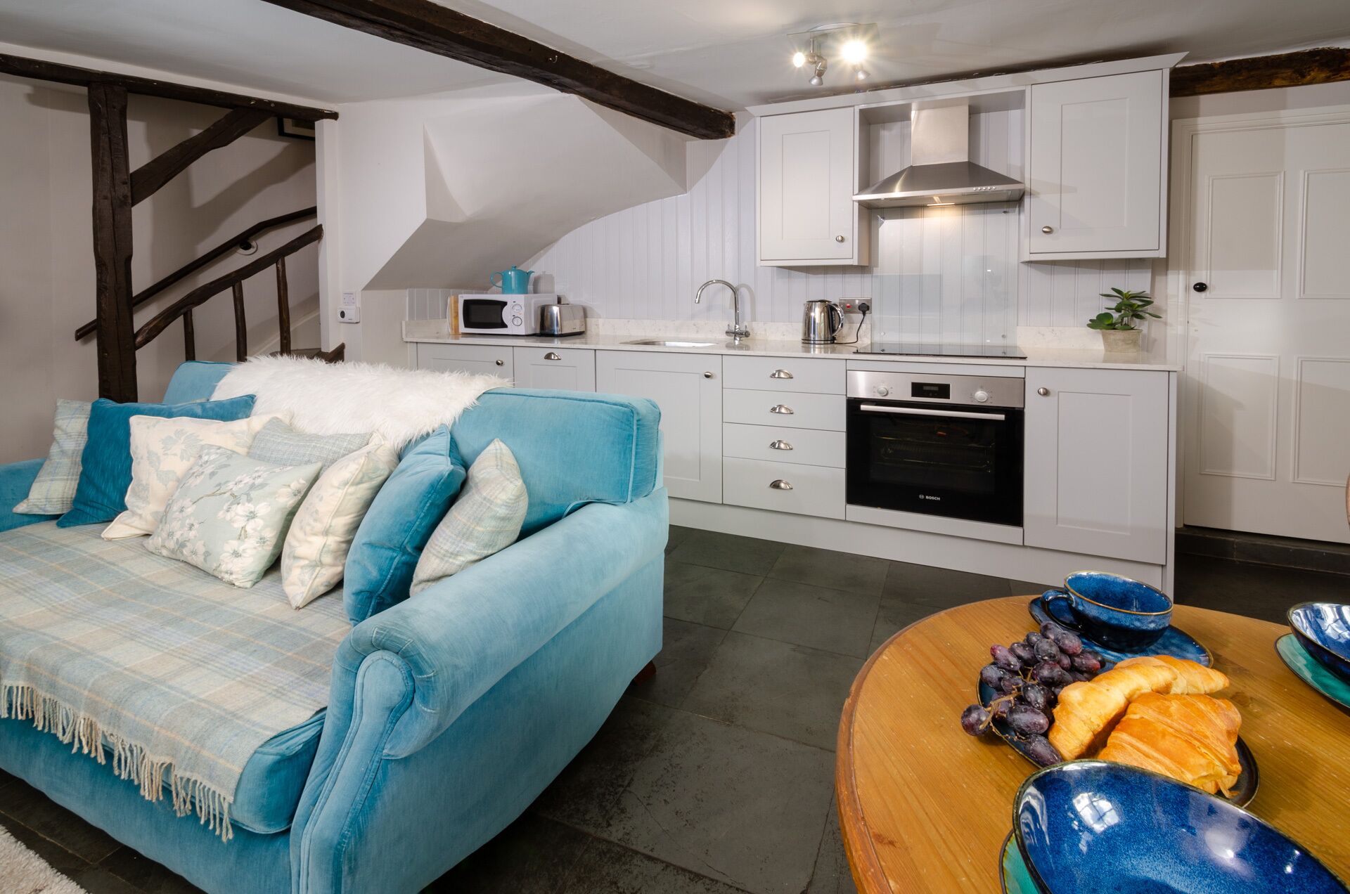 A modern kitchen with white cabinets, a Bosch oven, and a microwave. There's a blue sofa with a white throw blanket and blue cushions. A wooden table with a blue bowl and grapes is visible.