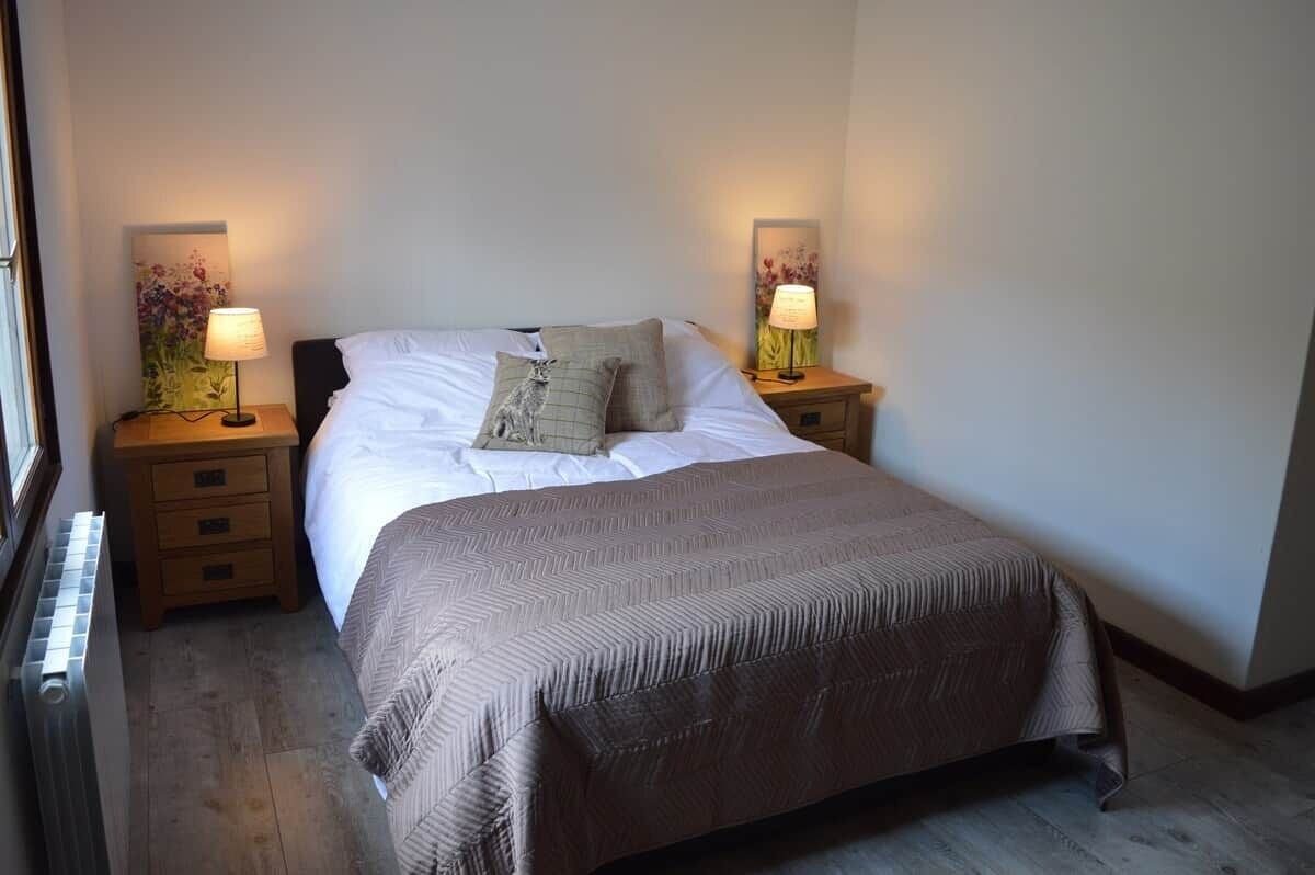 Una cama bien hecha con una colcha estampada, dos lámparas de noche y una mesita de noche con un cajón.