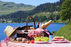 A picnic setup with a basket, wine, grapes, bread, and a plate of food by a lake.