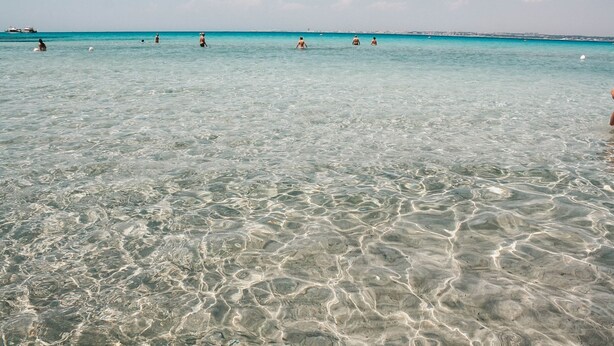 Een strand met helder ondiep water en verre bootjes.
