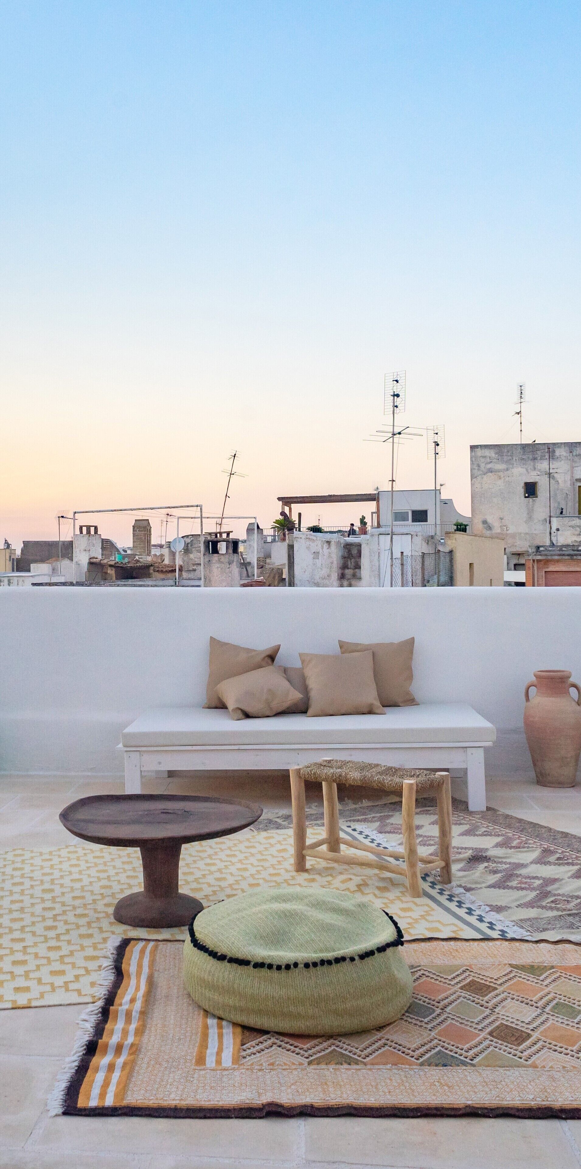 A rooftop terrace with a white sofa, a round table, and a woven pouf.