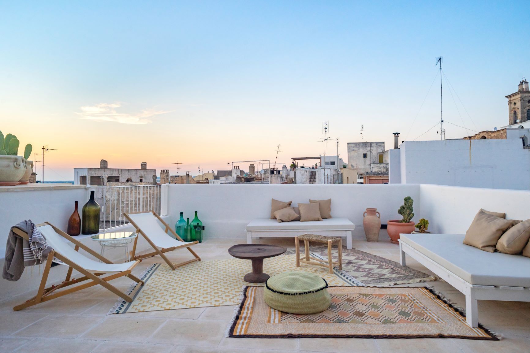 Eine Dachterrasse mit einem weißen Sofa, Lounge-Stühlen und einem kleinen Tisch. Überwiegend Kübelpflanzen und dekorative Flaschen.