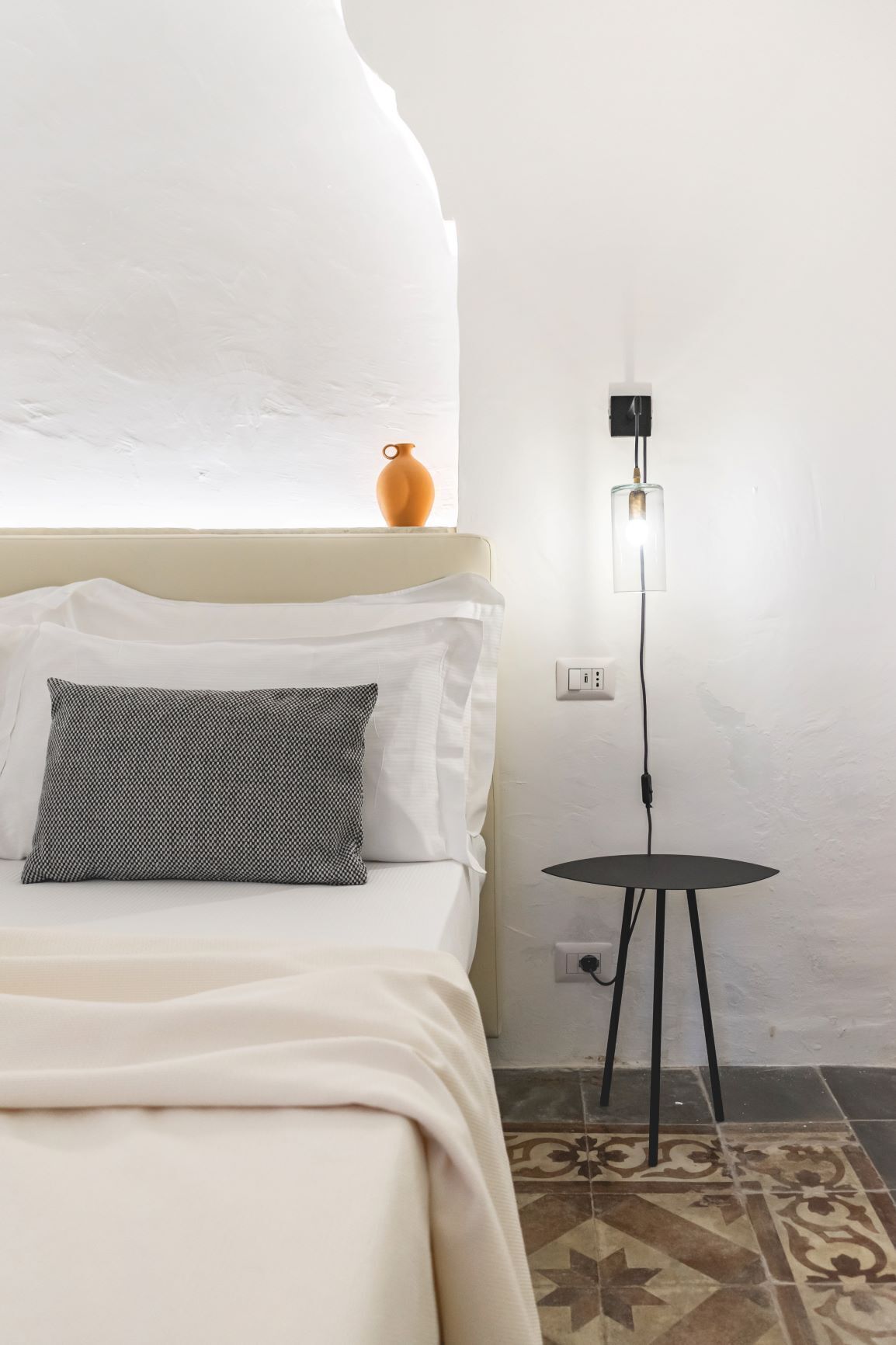 A bedroom with a bed, a small table, and a wall-mounted light fixture.