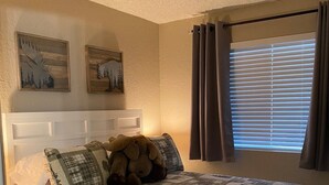 Ein Schlafzimmer mit Bett, Nachttisch, Fenster mit Vorhängen und zwei an der Wand hängenden Bildern.