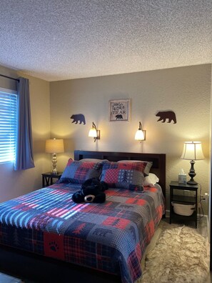 Une chambre à coucher avec un lit, deux lampes fixées au mur, une décoration murale sur le thème des ours et une fenêtre avec des rideaux.