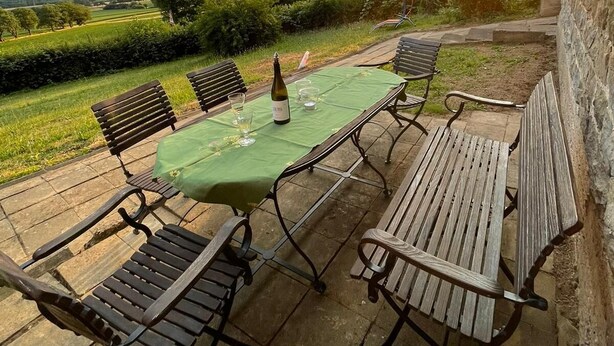 Un patio al aire libre con una mesa dispuesta para dos personas, rodeado de bancos de madera, con vistas a un paisaje pintoresco y una puesta de sol al fondo.
