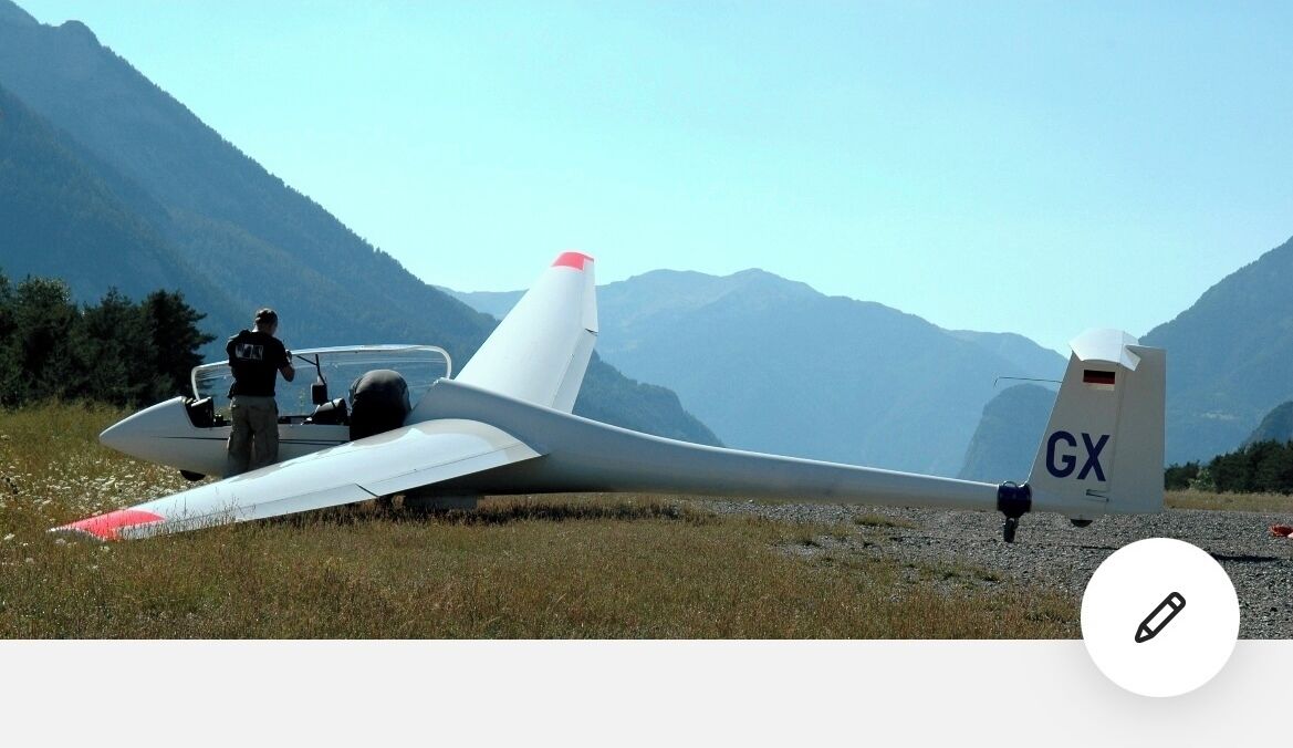 Un petit avion, immatriculé GX, est stationné sur un champ d’herbe, avec des montagnes en arrière-plan.