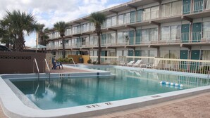 A swimming pool with a 9 FT 27 marker, surrounded by lounge chairs and palm trees, with a multi-story building in the background. - Cruise Port Inn (Cocoa)