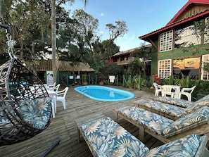 A wooden deck with a pool, lounge chairs, and a swing chair. - Pousada Canoa Azul (Garopaba)