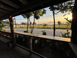 Vista desde un espacio al aire libre cubierto con un banco de madera, que da a una calle con valla y vegetación, incluyendo árboles y plantas.