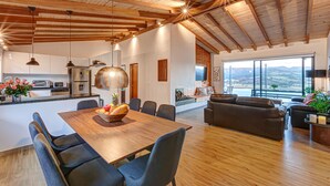 Une cuisine spacieuse équipée d’appareils modernes, un coin repas avec une table et des chaises en bois, et un salon avec un canapé et de grandes fenêtres offrant une vue sur l’extérieur.