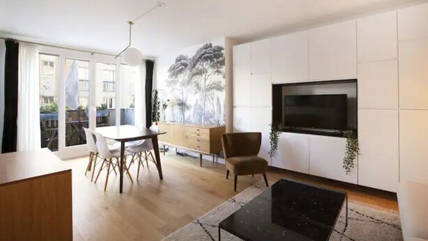 A modern living room with a dining area, a flat-screen TV mounted on a white wall, and a large window with a view of trees. - Havre de paix avec balcon (Paris)